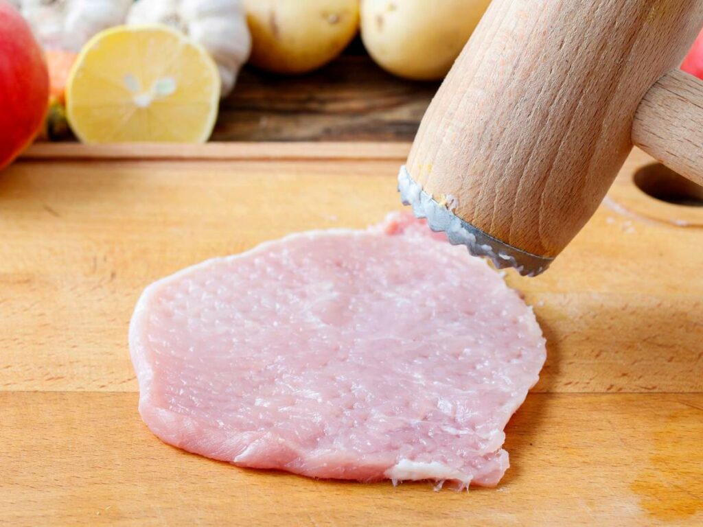 Raw pork cutlet being tenderized with a mallet on a wooden cutting board.