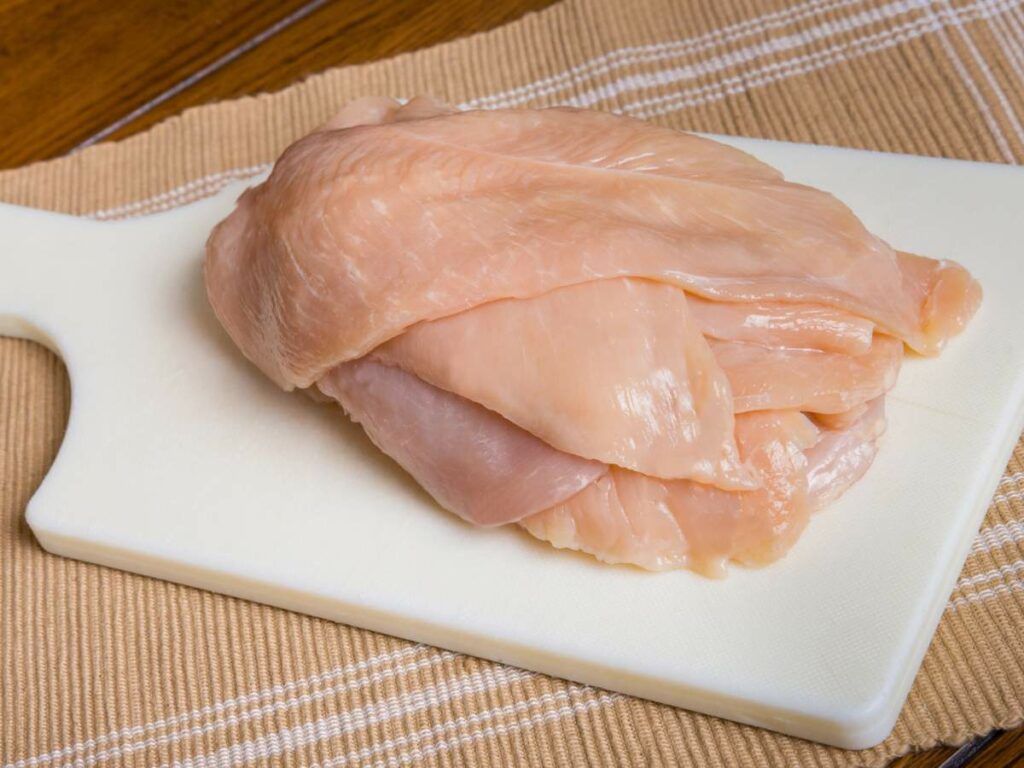 Raw chicken breast fillets stacked on a white cutting board over a brown striped cloth.