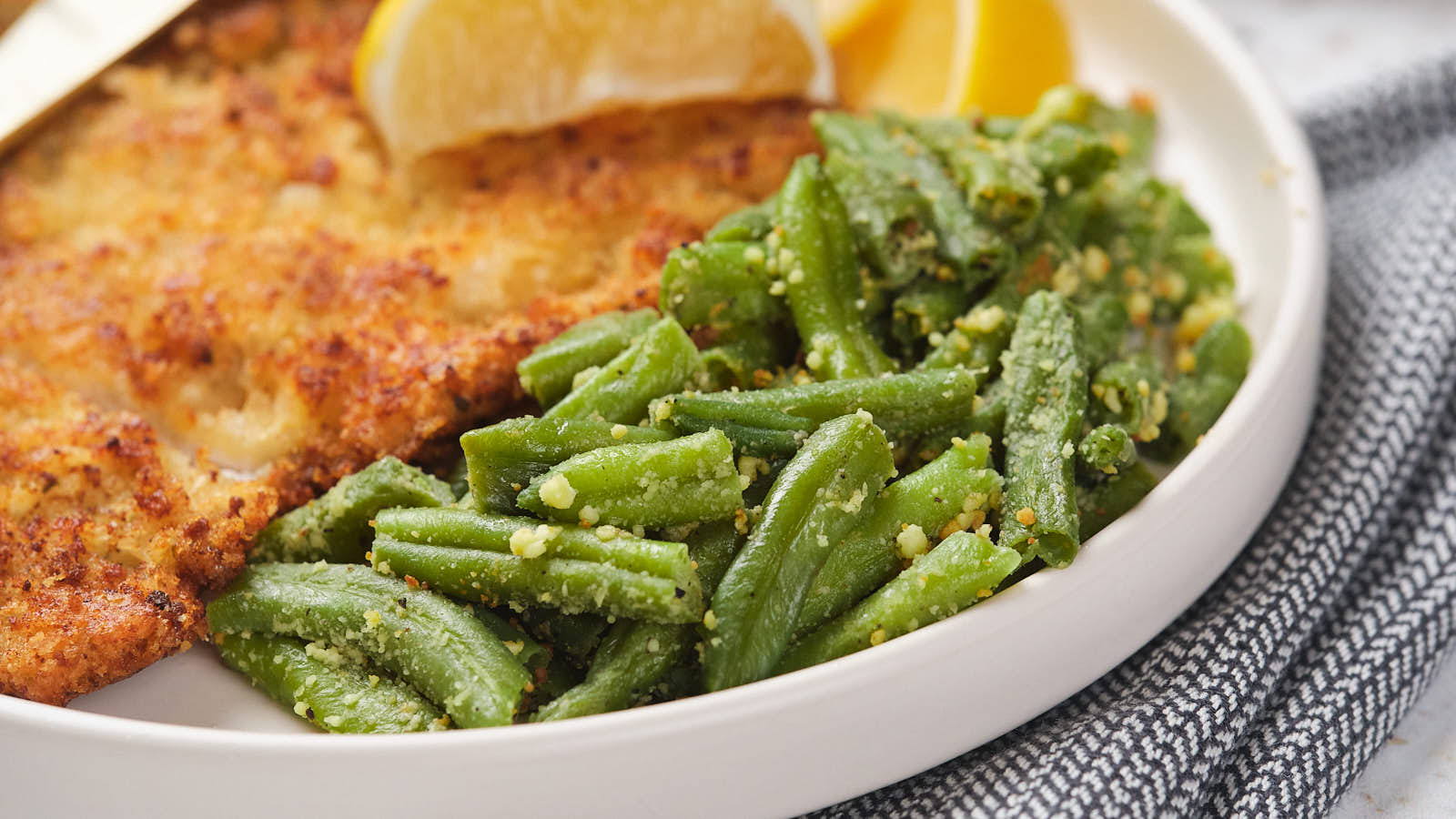 A plate with breaded chicken cutlet, roasted green beans with seasoning, and lemon wedges.