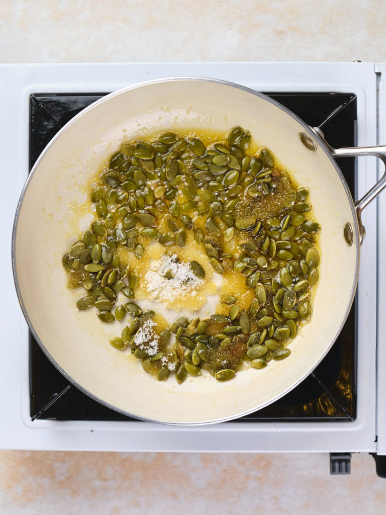 A frying pan with a mix of green pumpkin seeds, salt, and butter cooking on a stovetop.