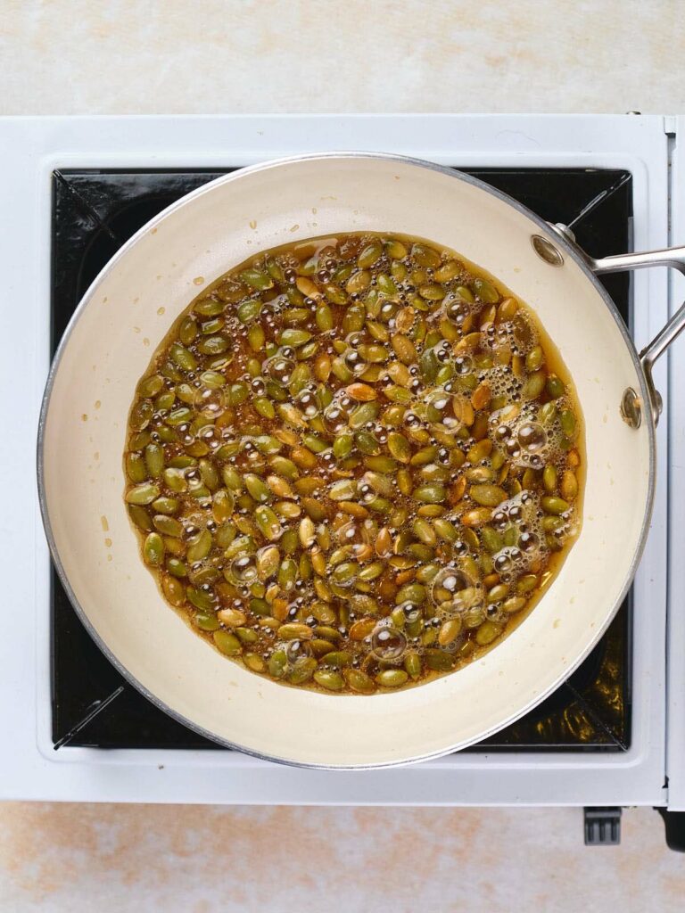 A sauté pan on a stovetop contains a mixture of caramelizing sugar and pumpkin seeds.