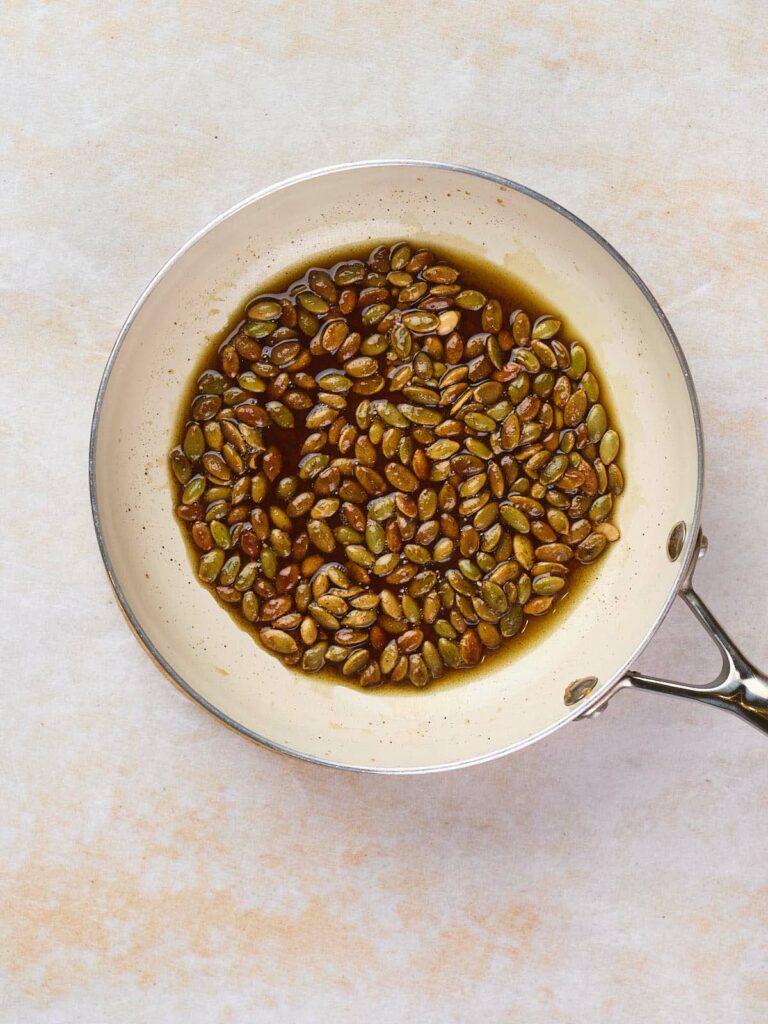 White frying pan filled with pumpkin seeds in oil, viewed from above on a light-colored surface.