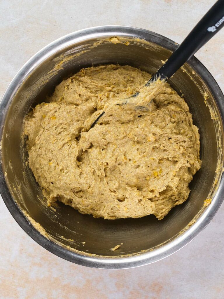 A stainless steel bowl filled with thick, beige-colored dough being mixed with a black spatula.