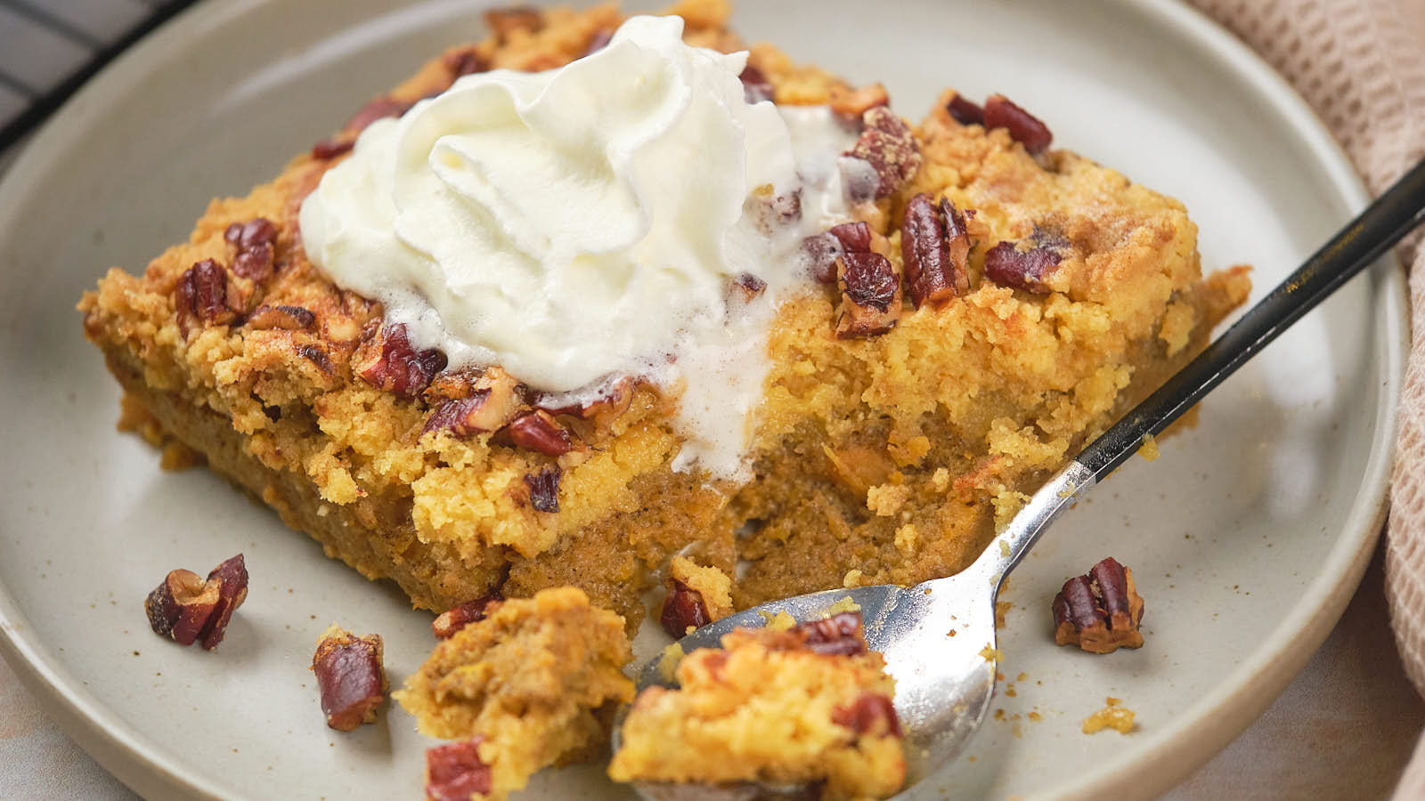 A slice of pumpkin dump cake topped with whipped cream and nuts on a white plate, with a spoon resting beside it.