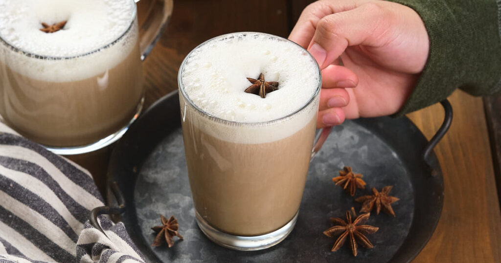 A hand holds a glass mug of frothy pumpkin spice chai latte topped with a star anise.