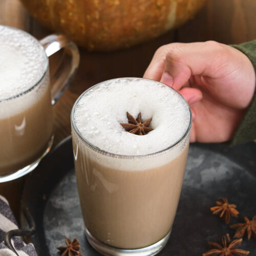 A hand holds a frothy pumpkin spice chai latte topped with a star anise.