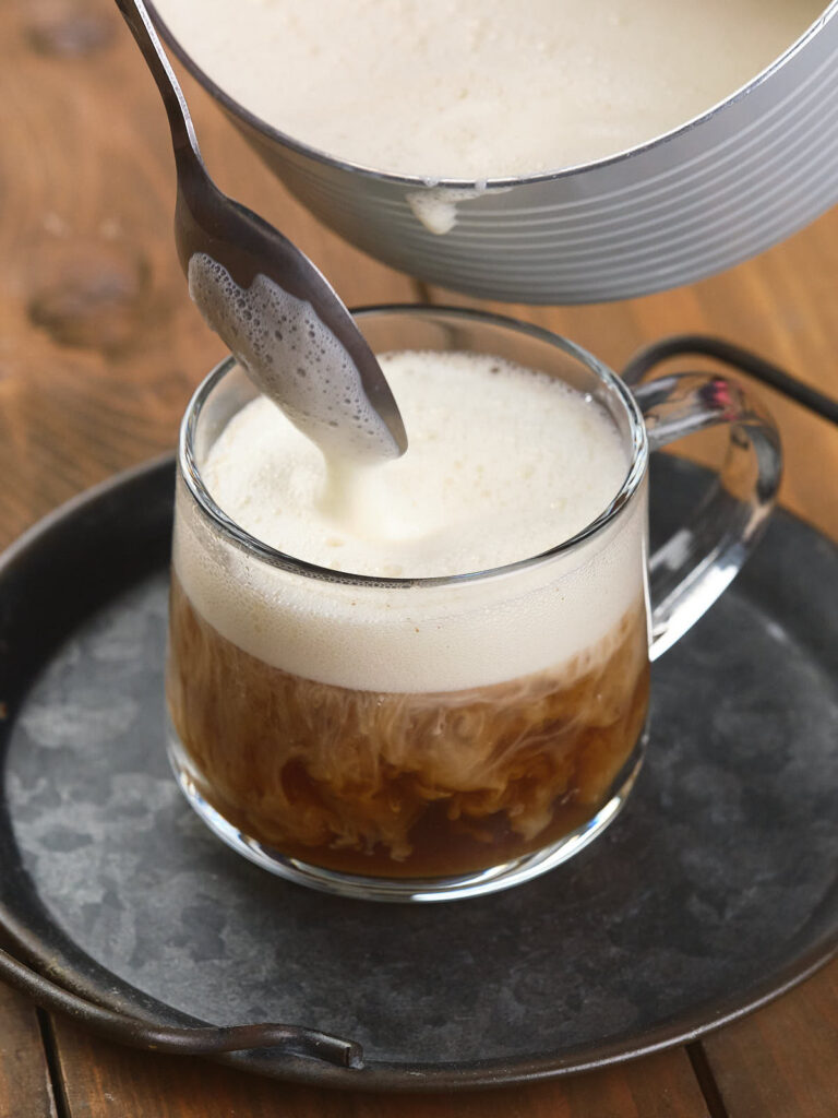 A glass mug filled with coffee and topped with foamy cream, on a round tray. A spoon is held above the mug, adding more foam.