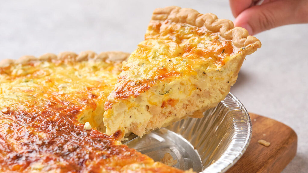 A hand lifting a slice of salmon quiche from a pie tin, revealing a golden, crispy top and creamy filling.