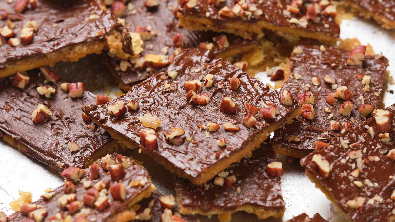 Close-up of saltine toffee squares topped with chocolate and chopped nuts on a silver surface.