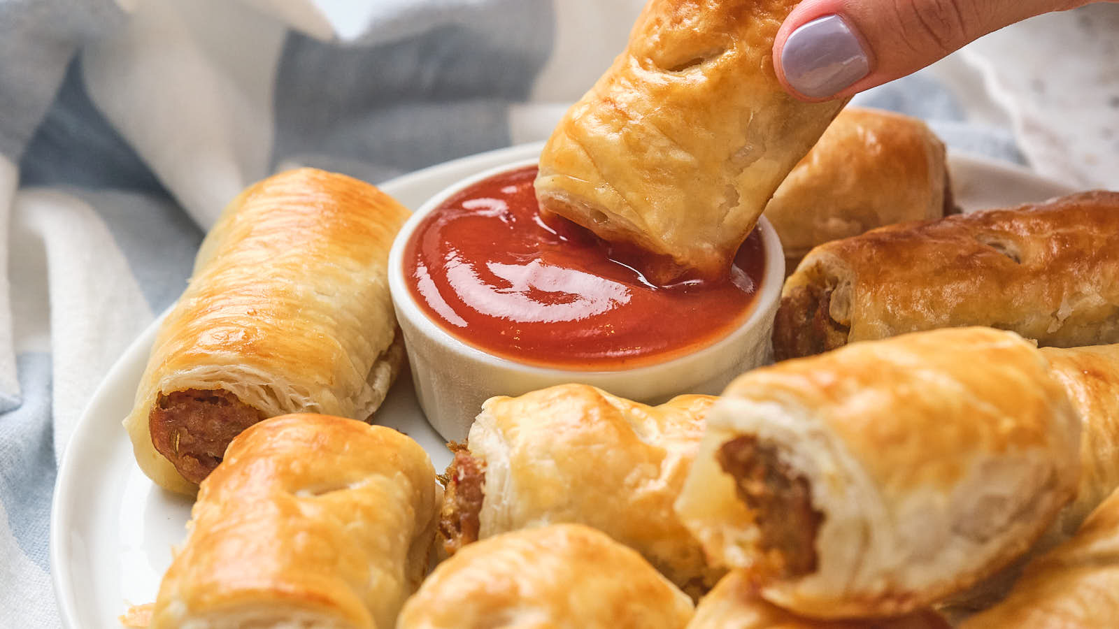 A hand dips a sausage roll into a dish of ketchup, surrounded by more sausage rolls on a white plate.