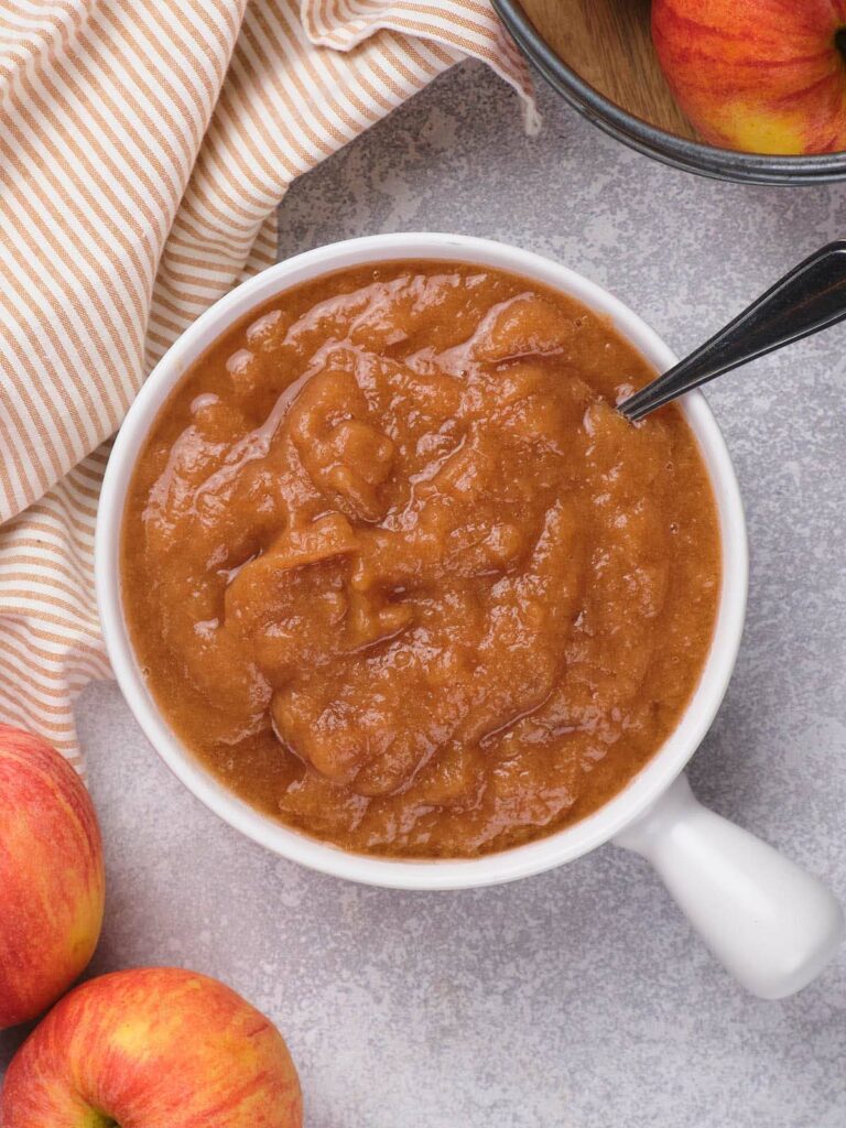 A white bowl of applesauce with a spoon, surrounded by fresh apples and a striped cloth on a gray surface.