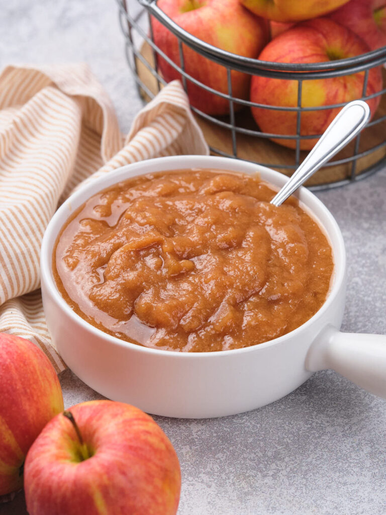 A white bowl of applesauce with a spoon, next to fresh apples and a striped cloth.