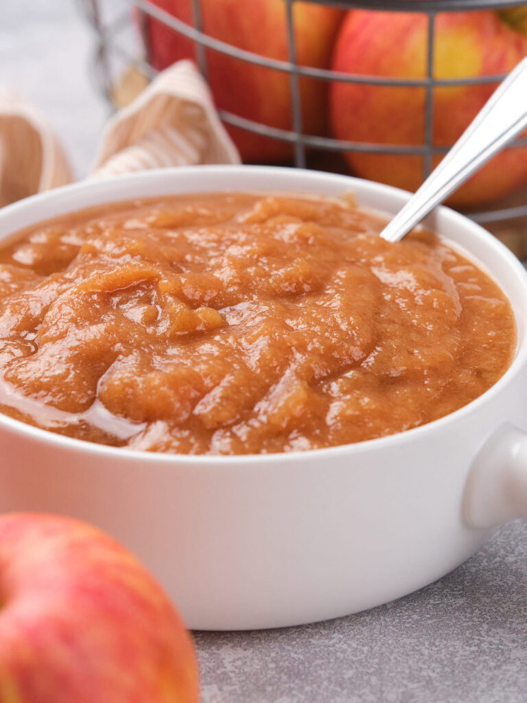 A bowl of applesauce with a spoon, set on a table with fresh apples and a striped cloth visible in the background.