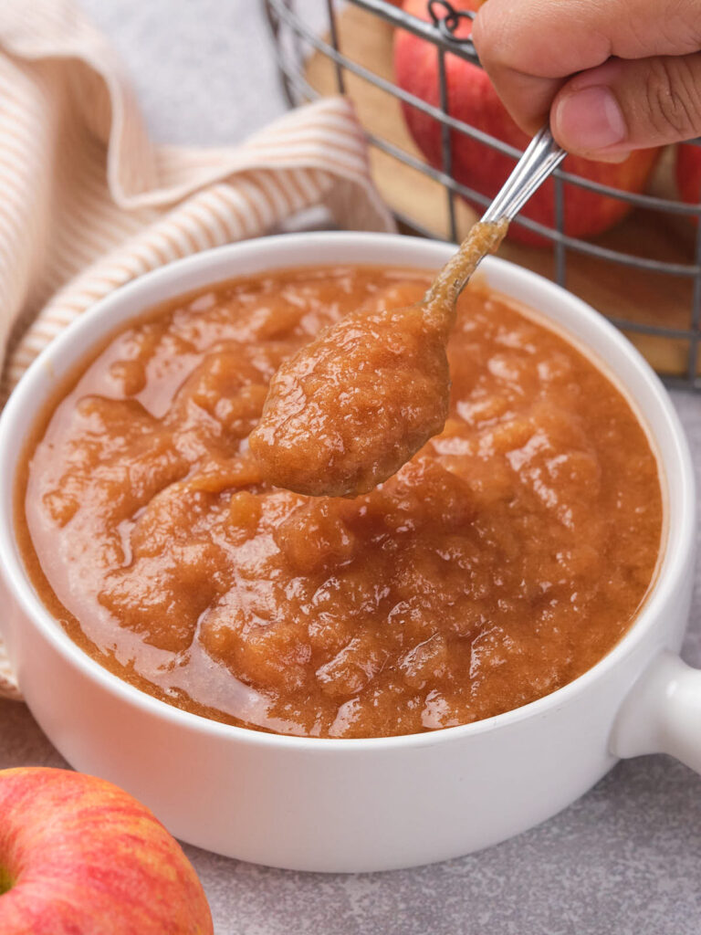 A hand holds a spoon above a bowl of applesauce. Nearby are apples and a striped cloth.