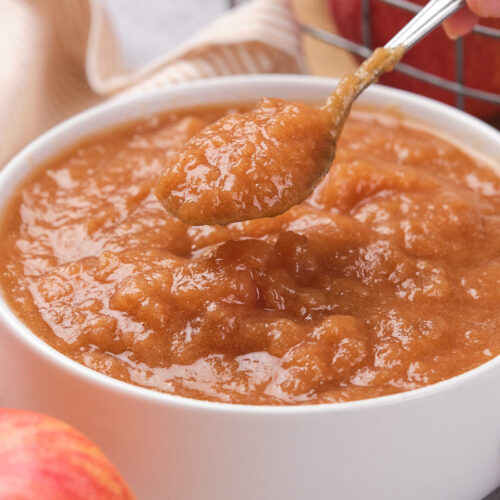 A spoonful of applesauce being lifted from a white bowl, with apples and a striped cloth in the background.