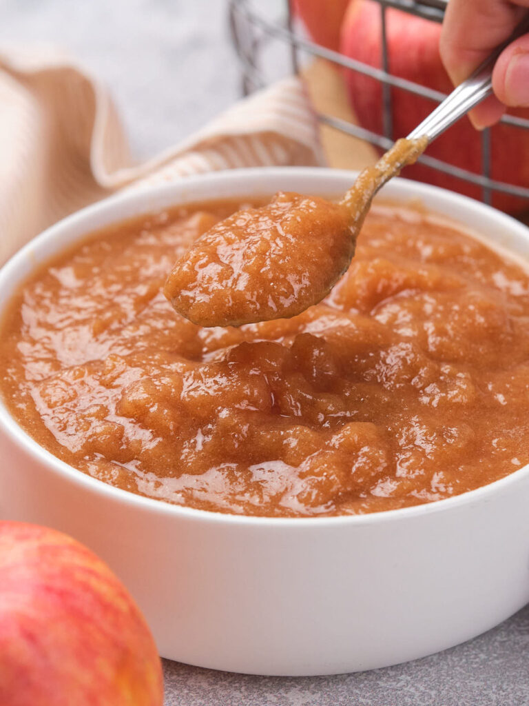 A spoonful of applesauce being lifted from a white bowl, with apples and a striped cloth in the background.