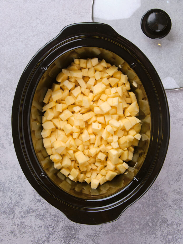 Chopped apples in a black slow cooker with a lid placed to the side.