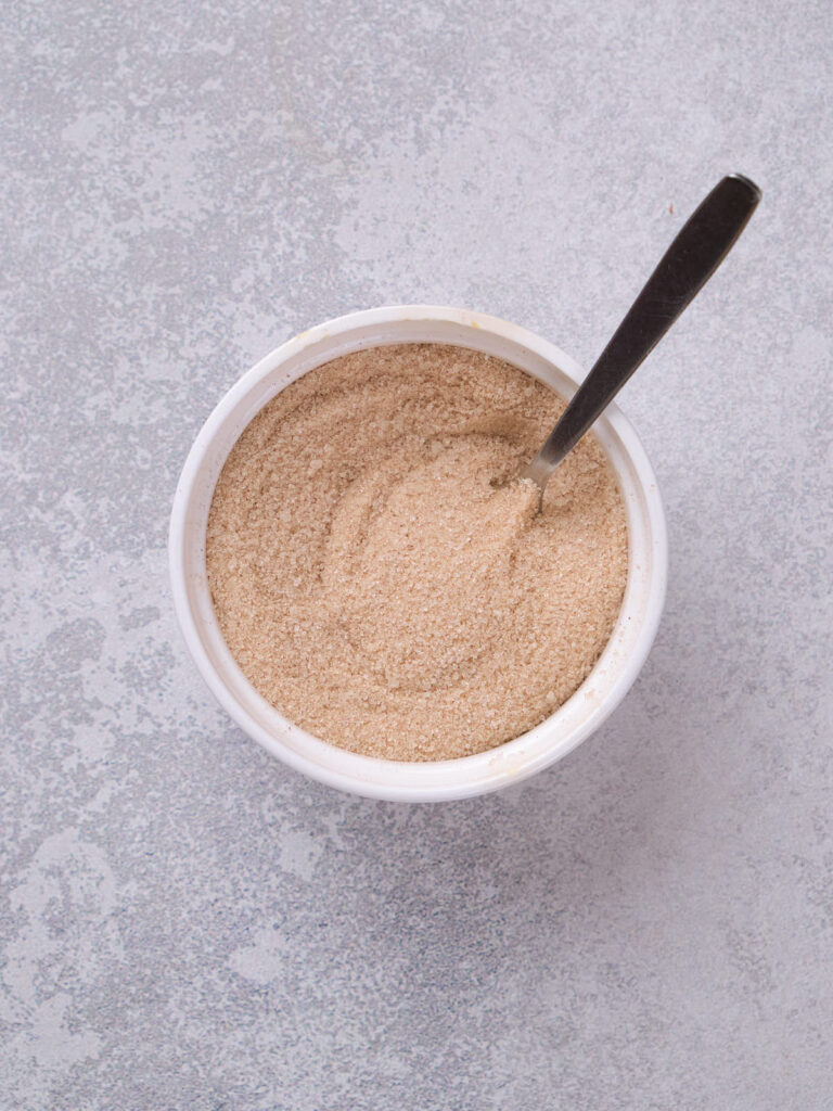 A bowl filled with a light brown powdered substance with a spoon inside, placed on a textured gray surface.