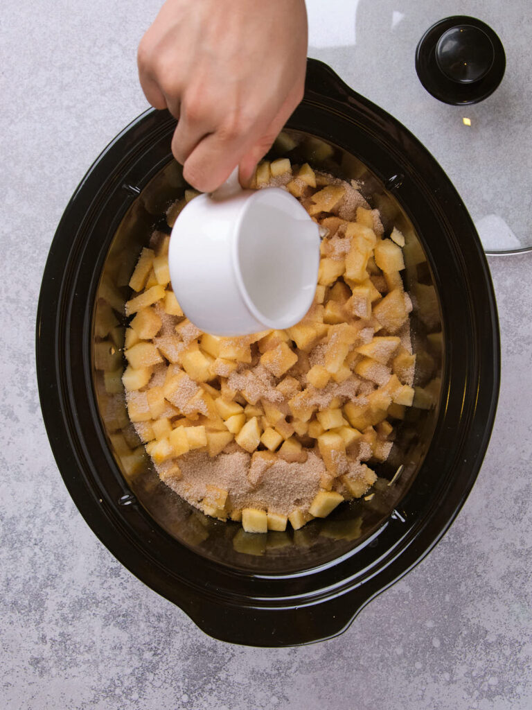 Hand pouring liquid into a slow cooker with chopped apples and cinnamon.