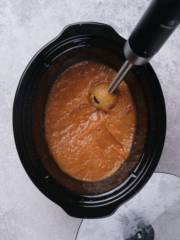 A black slow cooker with a hand blender mixing a thick orange soup or sauce on a gray surface.