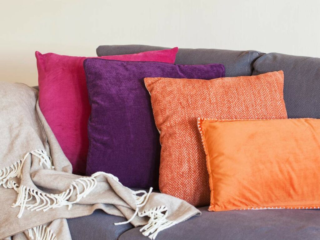 A gray sofa with a beige throw blanket and five colorful pillows.