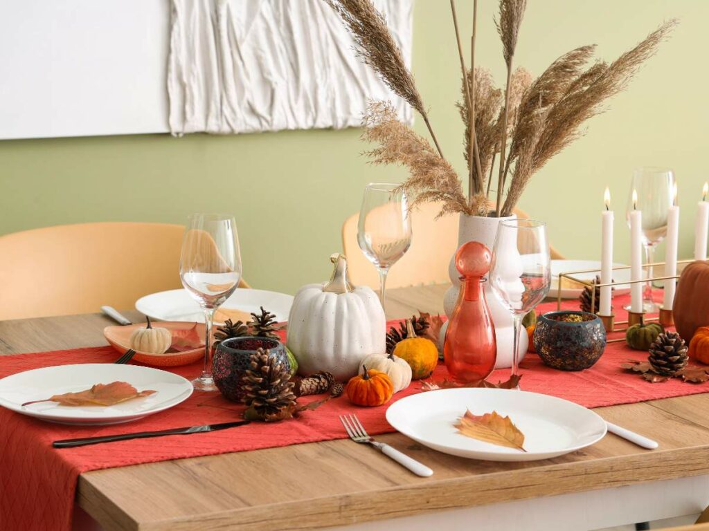 A dining table set with autumn-themed decorations, including small pumpkins, pine cones, candles, and dried grasses.