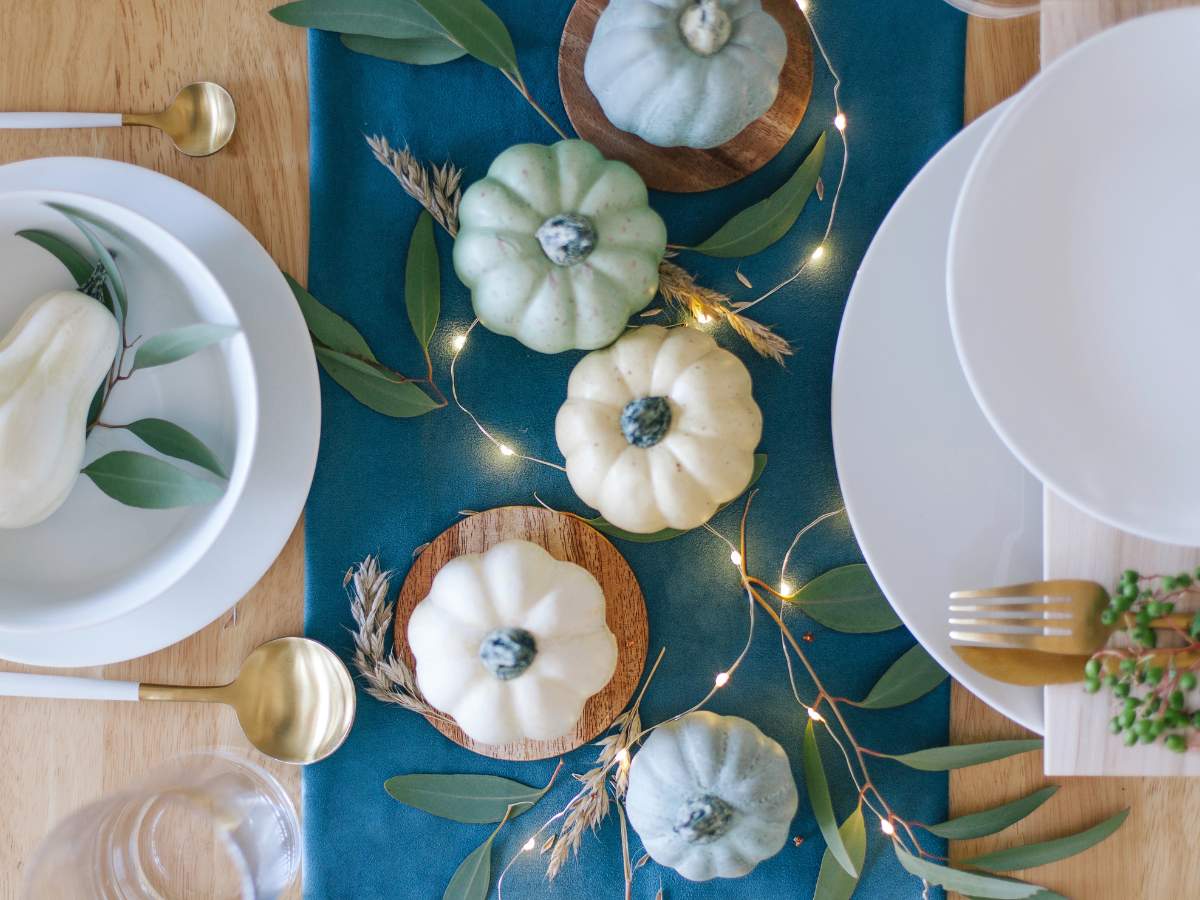 A table setting with white and green mini pumpkins on a teal runner, surrounded by leaves and string lights.