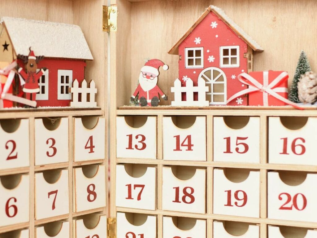 Wooden advent calendar with numbered drawers, featuring small red and white houses, a Santa figure, and a wrapped gift decoration on top.