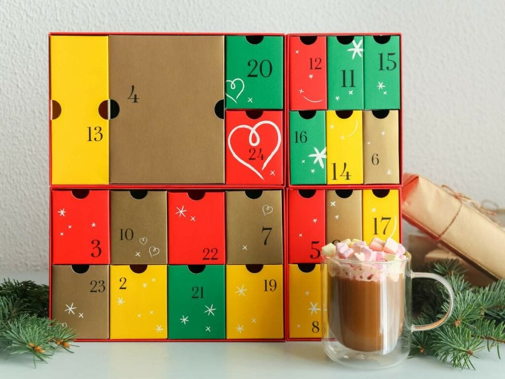 Festive advent calendar with numbered boxes in red, green, and gold, alongside a glass of hot chocolate topped with marshmallows..