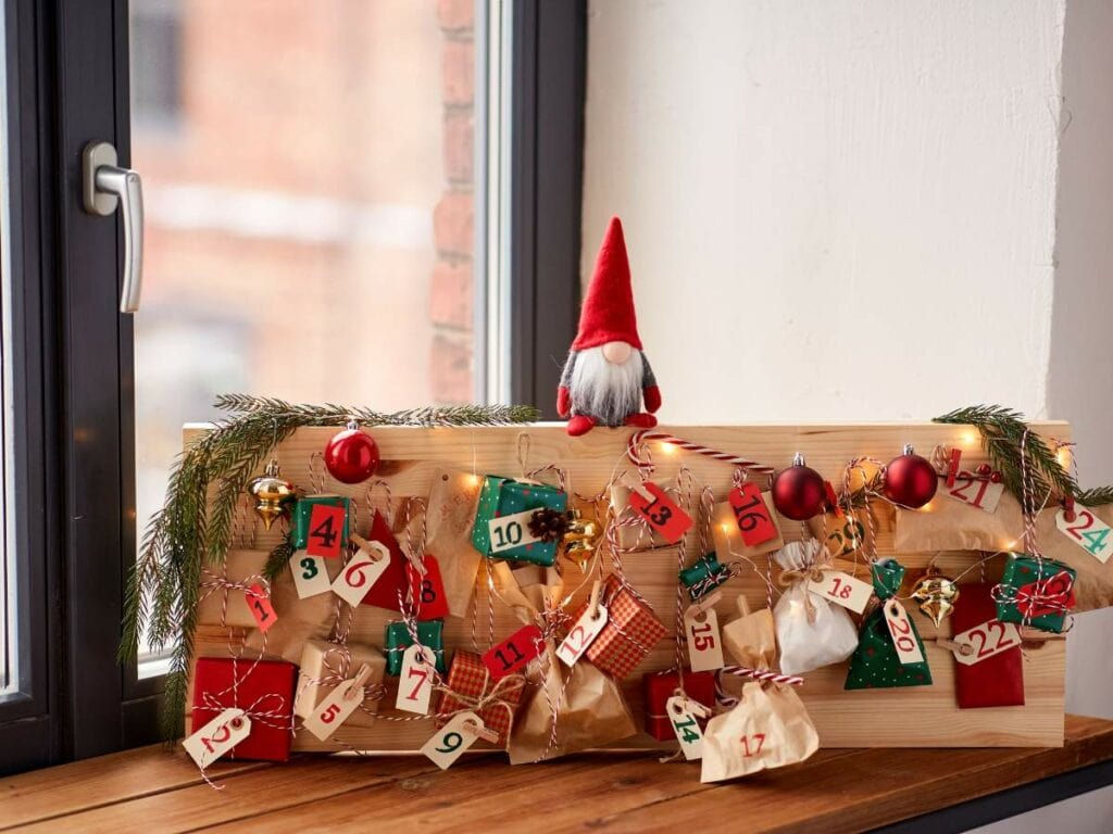 A homemade advent calendar on a windowsill features small numbered bags and ornaments, with a small gnome figure on top.