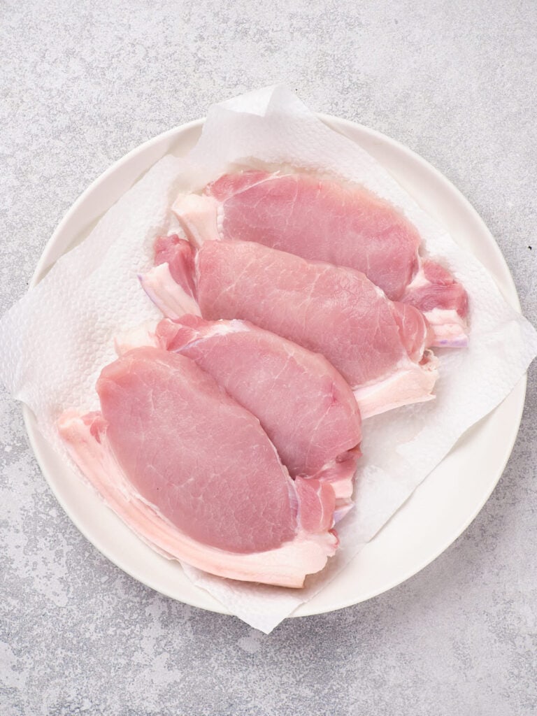 Four raw pork loin chops on a white plate lined with a paper towel.