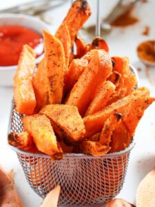 A basket of seasoned sweet potato fries with a bowl of ketchup in the background.