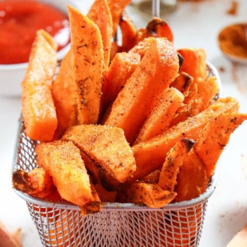 A basket of seasoned sweet potato fries with a bowl of ketchup in the background.