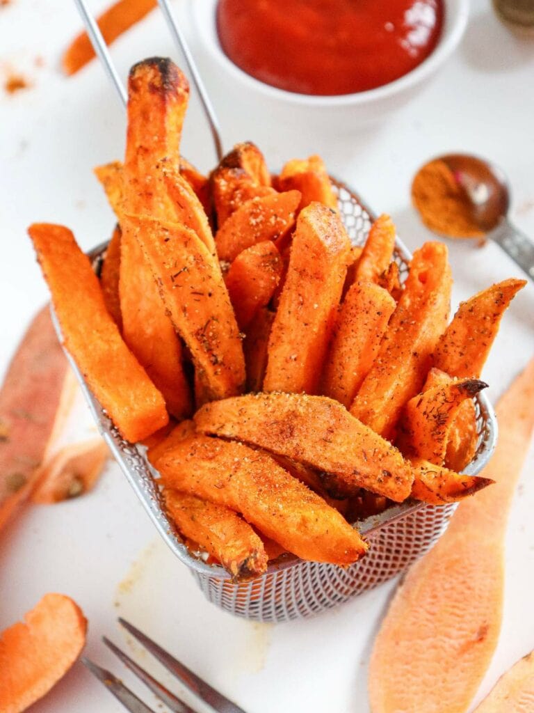 Basket of seasoned sweet potato fries with a bowl of ketchup in the background.