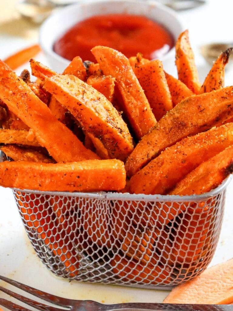 A basket of seasoned sweet potato fries with a bowl of ketchup in the background.