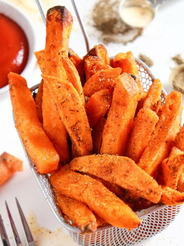 A basket of seasoned sweet potato fries on a white surface, accompanied by a small bowl of ketchup.