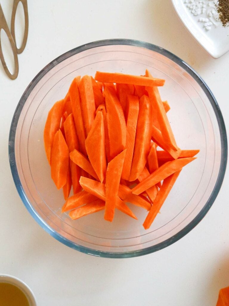 A glass bowl filled with orange carrot sticks on a white surface.