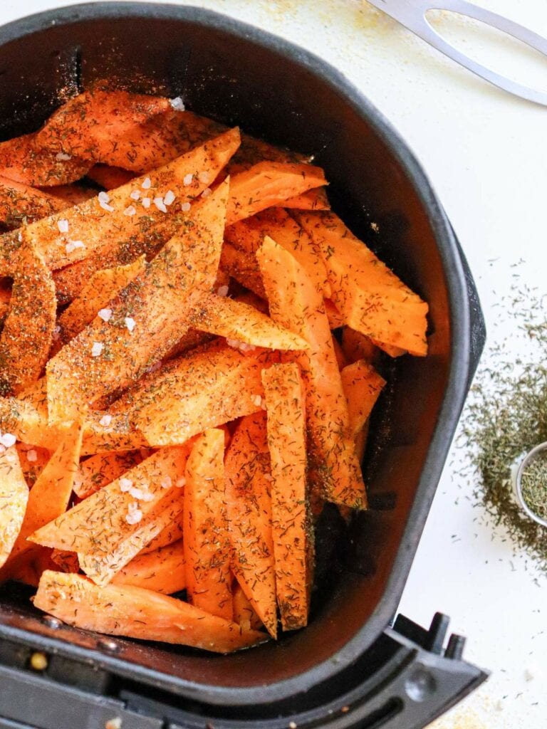 Sweet potato wedges seasoned with spices and herbs in an air fryer basket, with scattered herbs on the side.