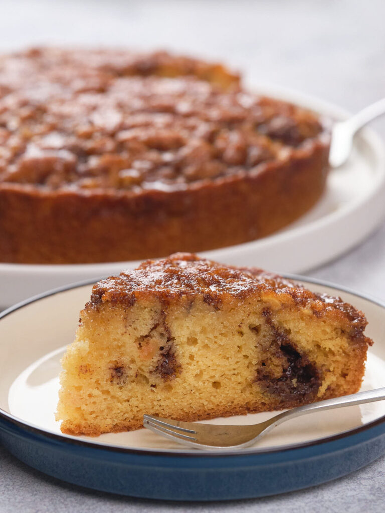 A slice of apple cinnamon cake on a plate with a fork, and the remaining cake in the background on a serving dish.