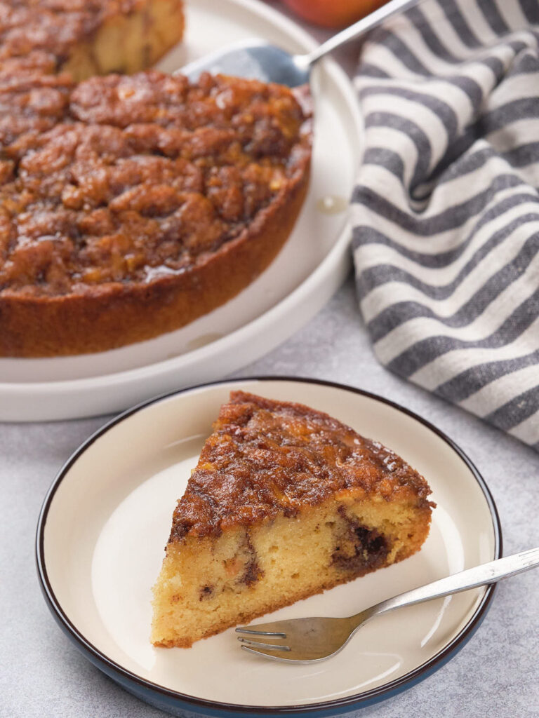 A slice of apple cinnamon cake on a plate with the whole cake in the background.