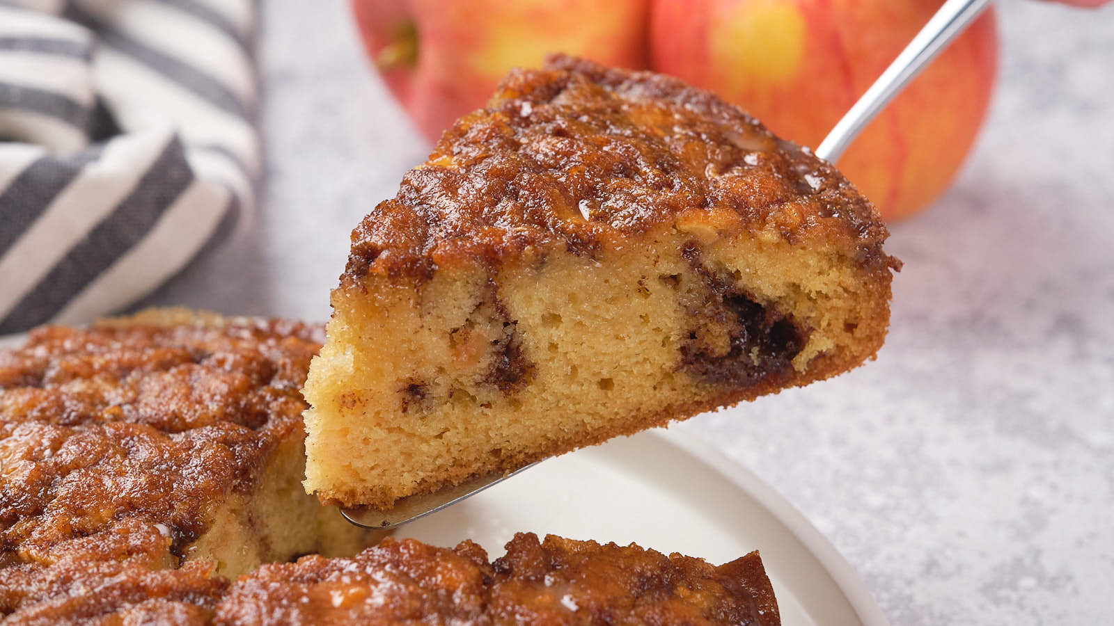 A slice of moist apple cinnamon cake with chocolate chunks is lifted from a whole cake.