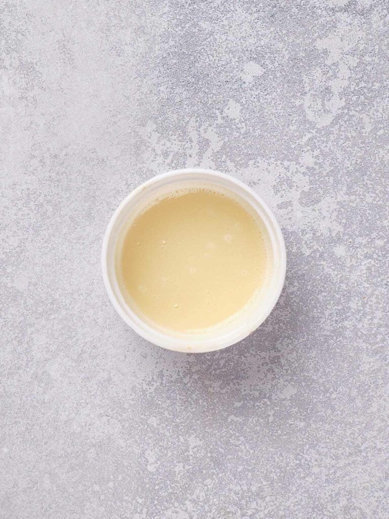 A small white bowl filled with a smooth, light beige liquid on a speckled gray surface.