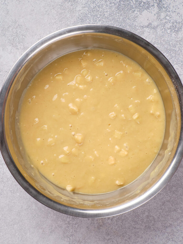 A metal bowl contains yellow batter with small chunks, resting on a light gray textured surface.