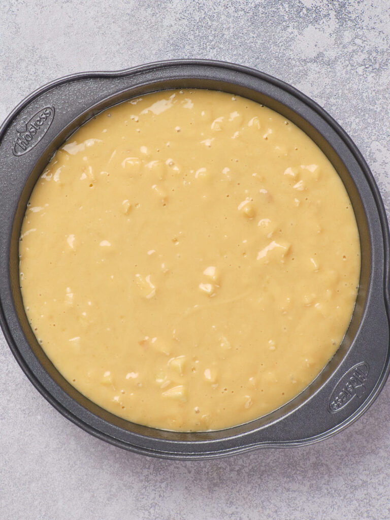 Cake batter with visible chunks poured into a round baking pan placed on a gray surface.