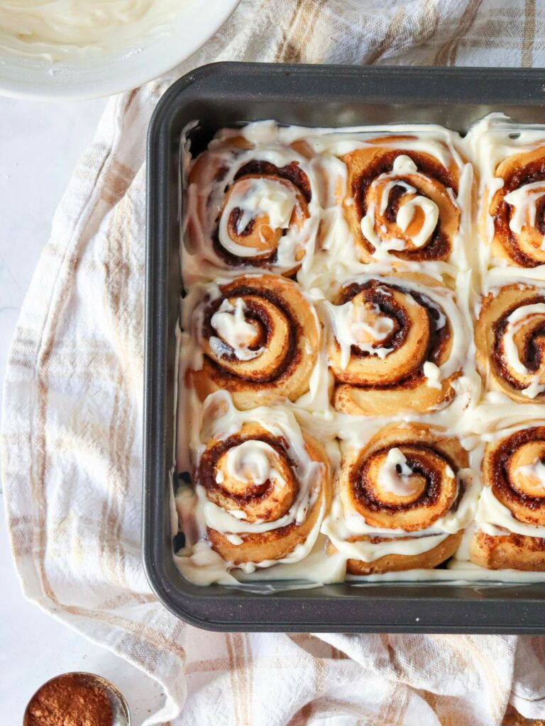 A baking tray with nine frosted cinnamon rolls on a striped cloth, next to a small dish of cinnamon.