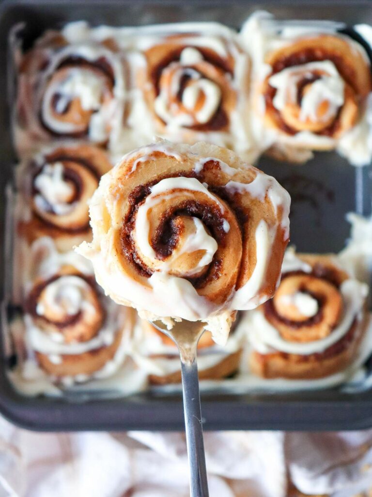 A close-up of a cinnamon roll with cream cheese icing on a spatula, above a pan of more cinnamon rolls.