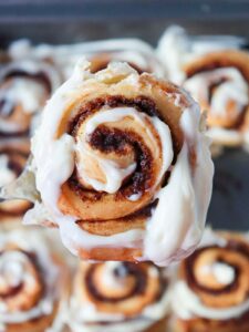 Close-up of a cinnamon roll with cream cheese frosting, held above a tray of similar rolls in the background.