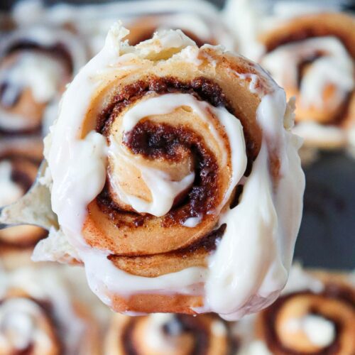 Close-up of a cinnamon roll with cream cheese frosting, held above a tray of similar rolls in the background.