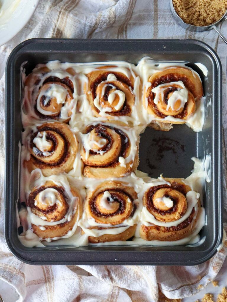 A tray of cinnamon rolls with cream cheese icing, one roll missing, on a checkered cloth with a bowl of brown sugar nearby.