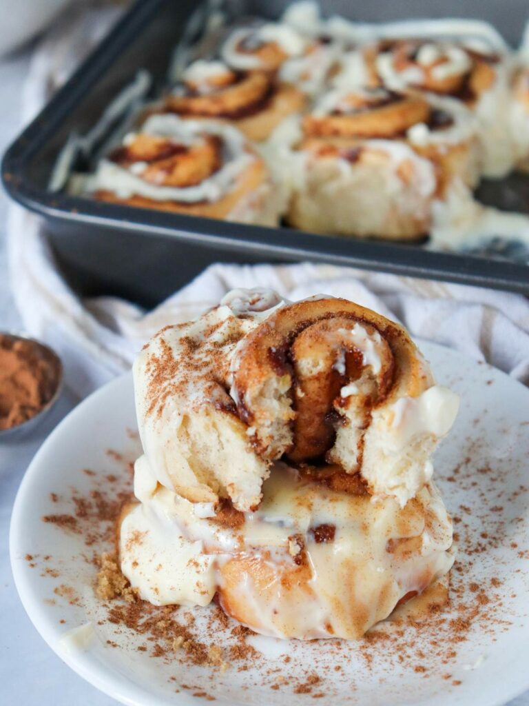 Close-up of cinnamon rolls on a plate, topped with icing and cinnamon, with a baking tray of rolls in the background.